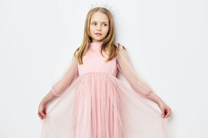 Young girl in a pink tulle dress posing elegantly with a crown, showcasing innocence and charm against a simple white background for creative portraits