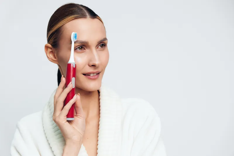 Young woman in white bathrobe holds electric toothbrush near face smiling gently isolated on white background personal hygiene oral care concept daily routine clean teeth