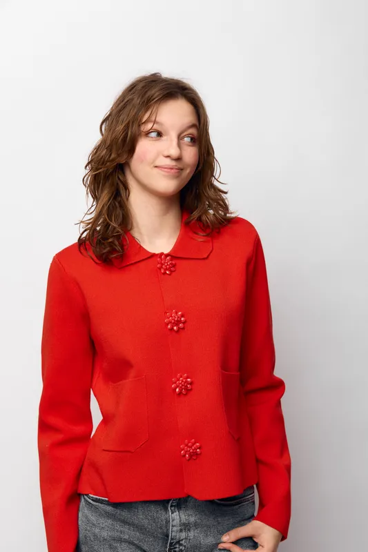 Young woman in red cardigan with decorative buttons looks sideways with slight smile. Casual outfit and natural hairstyle on plain white background. Concept of fashion, style, and confidence.
