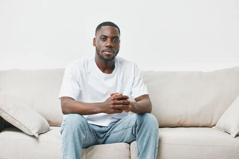 Casual portrait of a young Black man sitting on a beige couch, wearing a white t shirt and jeans, radiating confidence and calmness, ideal for lifestyle imagery