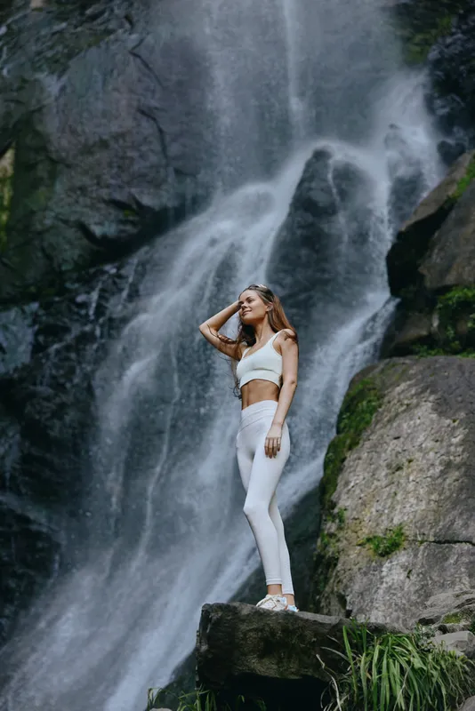 young woman in white activewear standing on rock near waterfall, fitness outdoors, natural beauty, summer adventure, energetic lifestyle