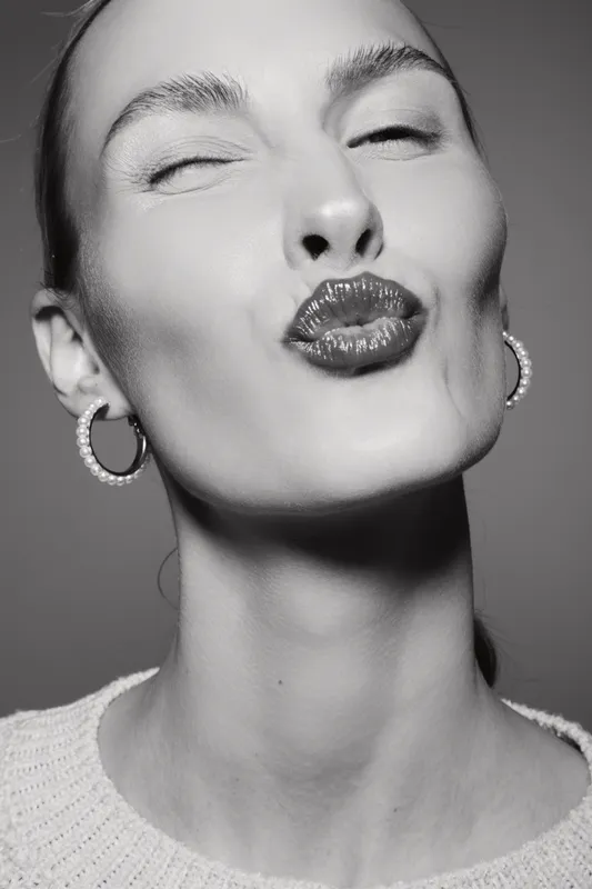 Closeup of a light skinned Caucasian woman with pearl earrings and red lipstick making a kiss face in black and white studio portrait.