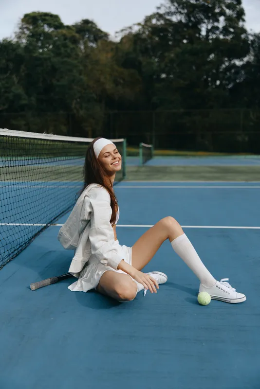 Young female tennis player in white sportswear smiling joyfully on the court, embodying sportiness and athleticism amidst a vibrant blue background with nature