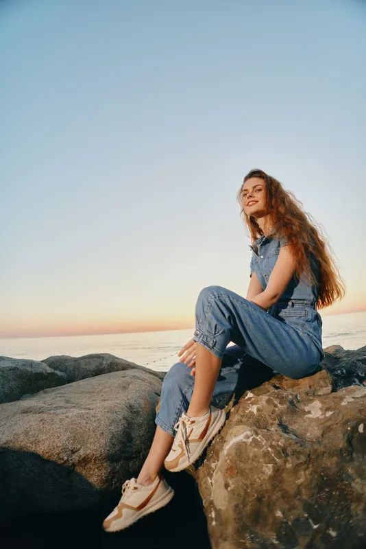 lifestyle woman in modern boho-western denim outfit sitting on rocks by the sea with film color tone, relaxed mood, casual street style, natural lighting