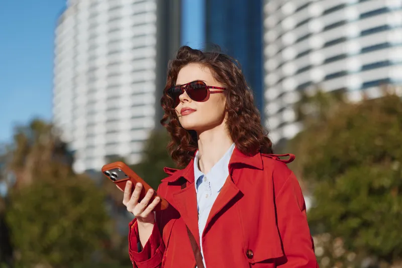 Stylish woman in a red coat holds a smartphone outdoors among modern buildings, looking confident and focused as she checks a message in bright daylight.