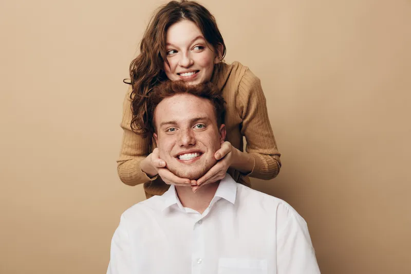 Happy young couple poses together on beige background. Woman in brown sweater holds man’s face gently while both smile. Concept of love, friendship, and positive emotions.