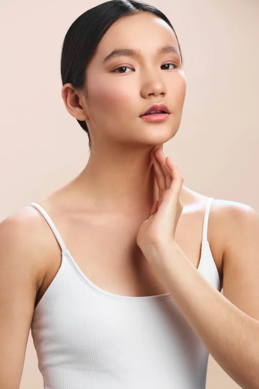 asian woman in soft studio lighting poses gracefully, showing clear skin and subtle makeup, exuding calm confidence and elegance as she rests her hand near her neck.