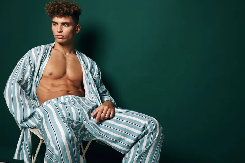 Young male model with curly hair wearing striped suit without shirt sitting on stool in studio with dark green background. Casual style pose and fashion with serious expression and relaxed hands.