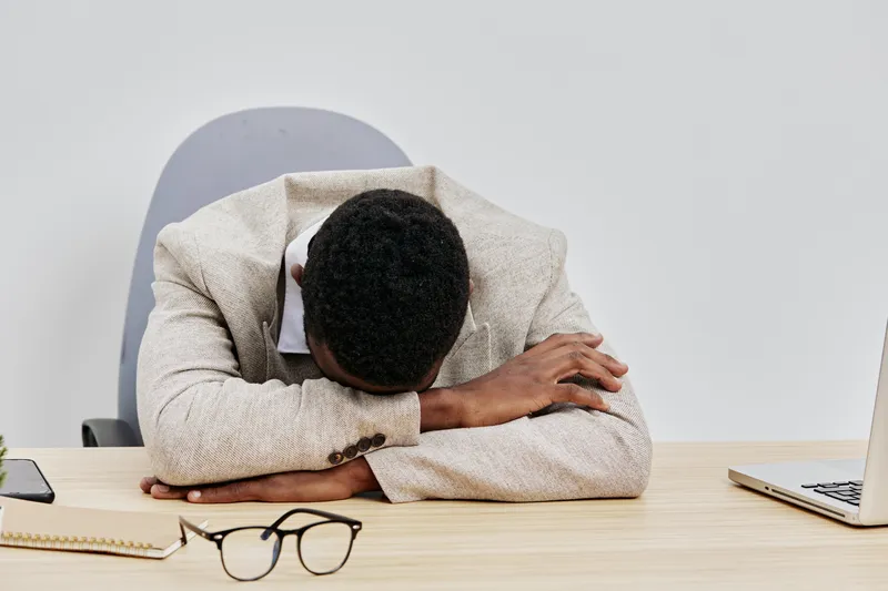 Professional young man resting his head on folded arms at a desk with laptop and glasses, exhausted or stressed, indoor office setting, neutral background, casual business attire, concept of fatigue o