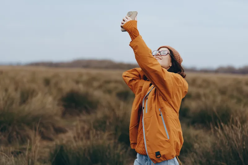Woman lifestyle outdoors in autumn field, wearing an orange jacket and knit hat, standing in tall grass with raised arm and glasses, calm nature.