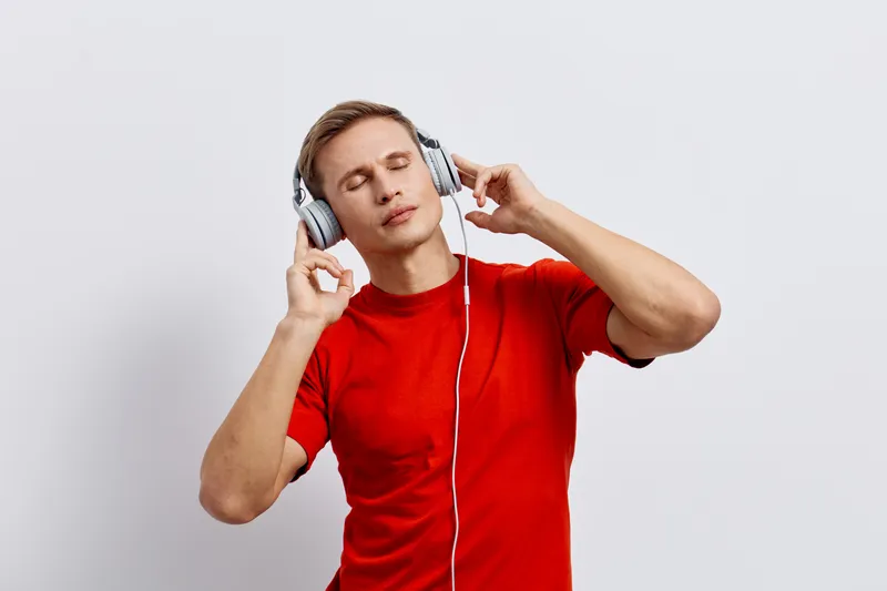 Young caucasian man in red t-shirt listen music with headphones closed eyes enjoy relaxed lifestyle concept mental health and gadgets isolated white background