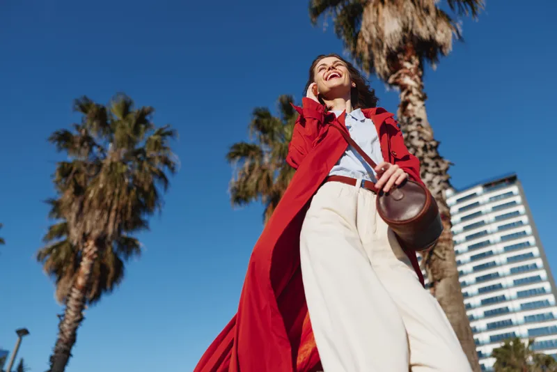 A young woman wearing a red coat and white pants smiles joyfully while walking outdoors on a sunny day with palm trees and a building in the background.