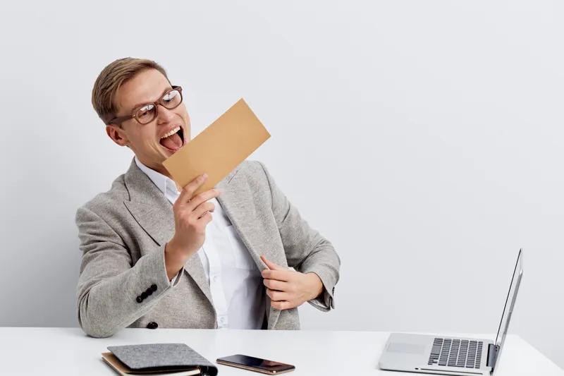 Professional young businessman in gray blazer and glasses holding a greeting card, expressing excitement and joy, sitting at a desk with a laptop and smartphone, isolated on plain white background. Bu
