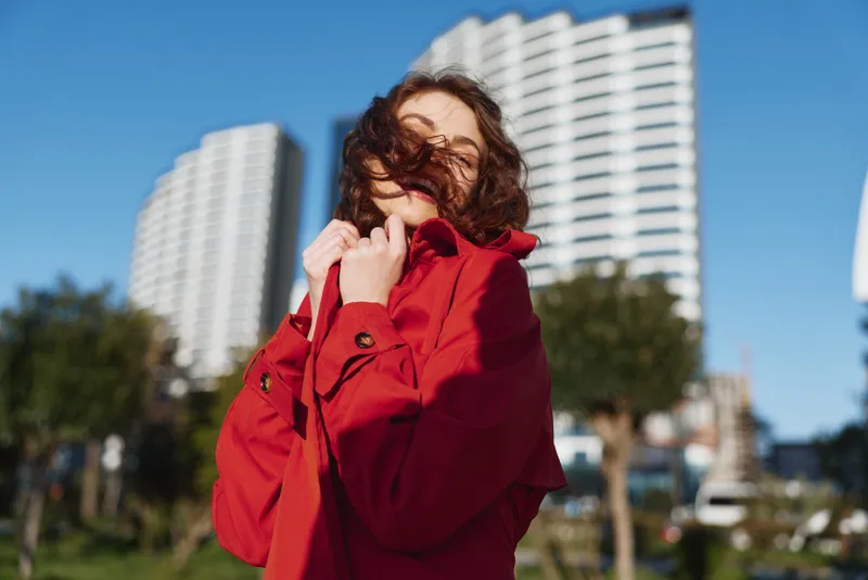 Young woman with curly hair wears a bright red coat and smiles gently while standing outdoors with tall buildings blurred in the background on a sunny day.