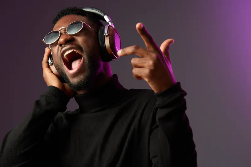 Excited young Black man singing with headphones, wearing sunglasses against a vibrant purple background Music, joy, and energy are highlighted
