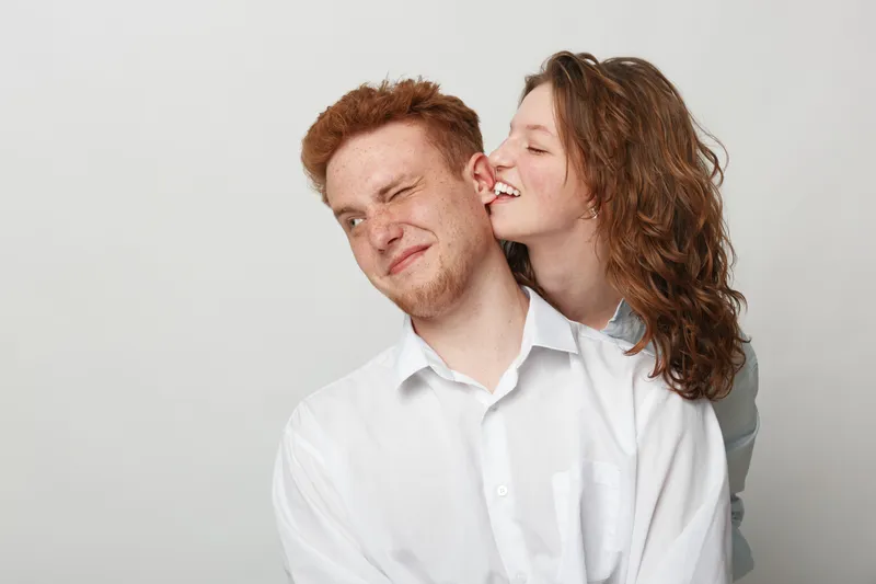 Young couple in white shirts share playful moment, woman whispering in man’s ear with smile. Studio portrait on light background, concept of love, intimacy, and happiness.