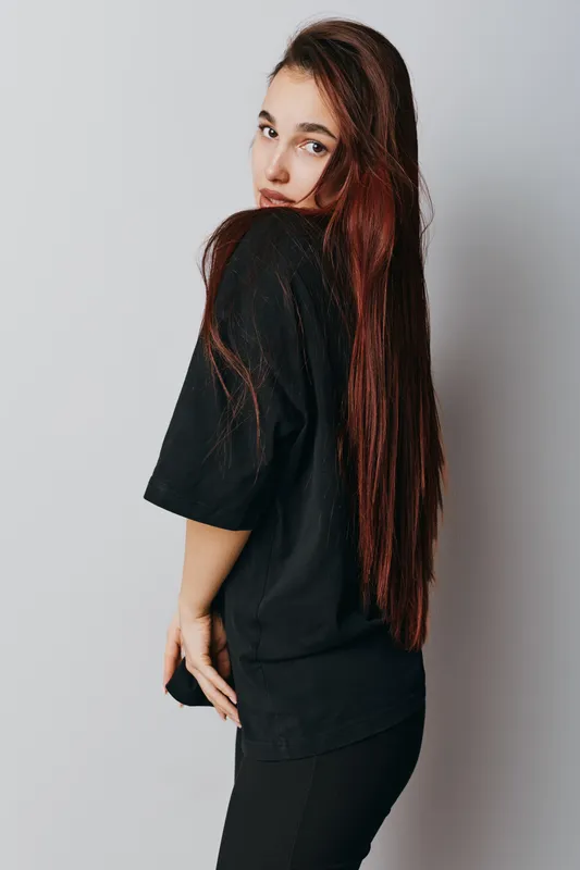 Young woman with long straight hair wearing black casual t shirt and leggings posing sideways against white background in studio. Portrait of female model with natural makeup and relaxed expression.