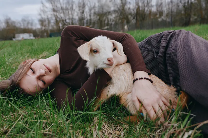 Cheerful lifestyle moment outdoors, woman lying on grass embracing a young goat kid, warm clothing, soft sunlight, gentle smile, peaceful connection, natural colors, rustic simplicity
