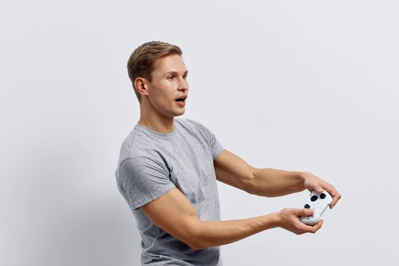 Young surprised man wearing casual gray t-shirt holding a small remote control device with white and black accents isolated on plain white background studio portrait. People lifestyle concept