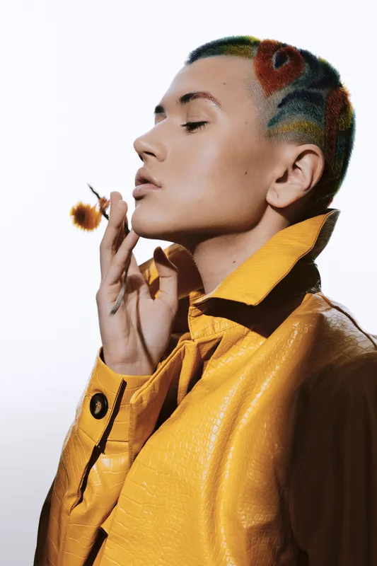 woman with colorful shaved hairstyle wearing a vibrant yellow leather jacket, posing thoughtfully with closed eyes against a simple white background, stylish and modern fashion