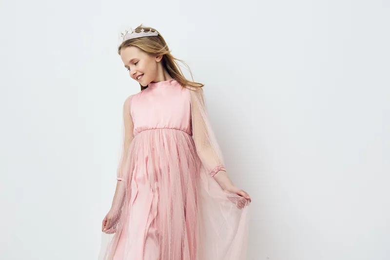 Joyful girl in a pink princess dress with a crown, displaying happiness against a soft white background, ideal for themes of childhood dreams and celebrations