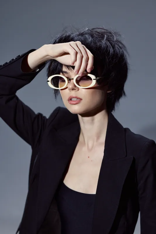 Stylish woman with short hair wearing unique round sunglasses and black outfit posing confidently in studio lighting.