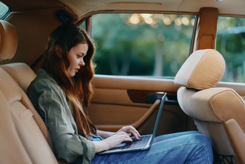 Young woman working on a laptop in a vintage car's backseat, showcasing a relaxed and focused attitude amidst a natural green background.
