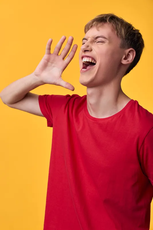 Energetic youth in a red shirt shouts with hand cupped near the mouth against a bright yellow background, capturing bold expression, loud voice, and attention grabbing action