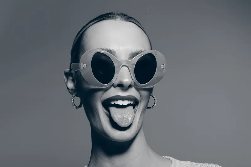 Young white woman with fair skin and slicked back hair wearing round sunglasses sticks out tongue in playful studio portrait.