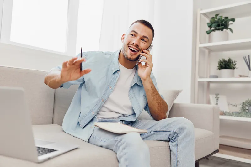 Smiling man engaged in a cheerful phone conversation, casually dressed in light blue attire, surrounded by a modern living space with natural light.