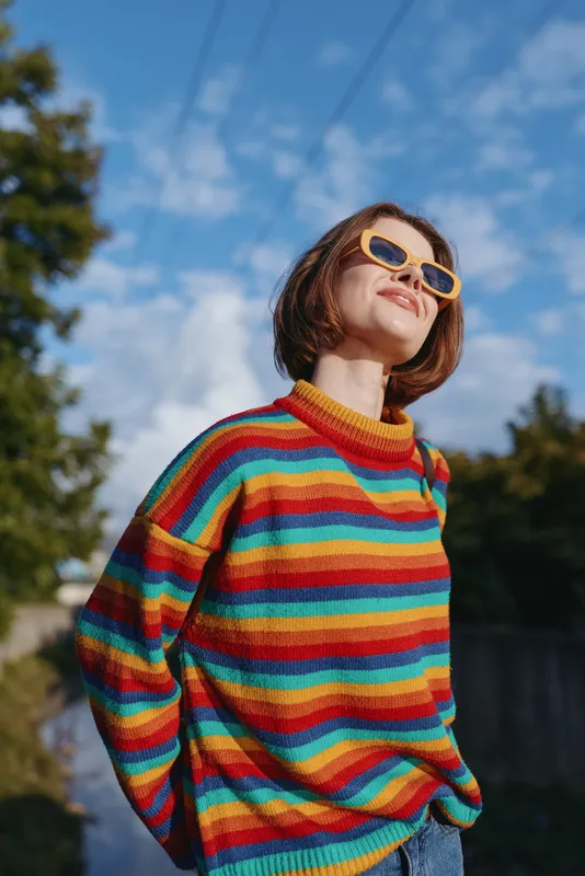 Woman smiling in a colorful striped sweater and yellow sunglasses, short hair bob outdoors under blue sky. Young adult in casual outfit looking up, sunny joyful lifestyle portrait and travel vibe.