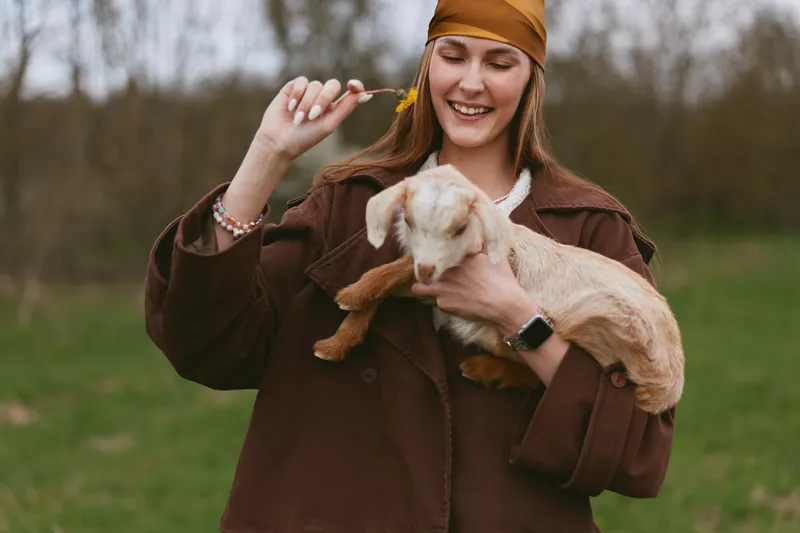 A smiling woman outdoors holds a goat kid, wearing a brown jacket and headband. A candid lifestyle moment showing gentle bonding with a small animal in a grassy countryside setting.