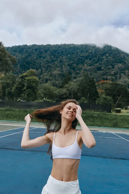 Young woman enjoying a joyful moment on a tennis court, wearing a white crop top and skirt, surrounded by lush greenery, capturing the essence of a vibrant, active lifestyle