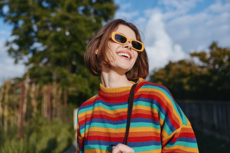 Woman smiling outdoors in a colorful striped sweater and yellow sunglasses, short hair bob and casual outfit in a sunny park, joyful travel lifestyle portrait with carefree mood.