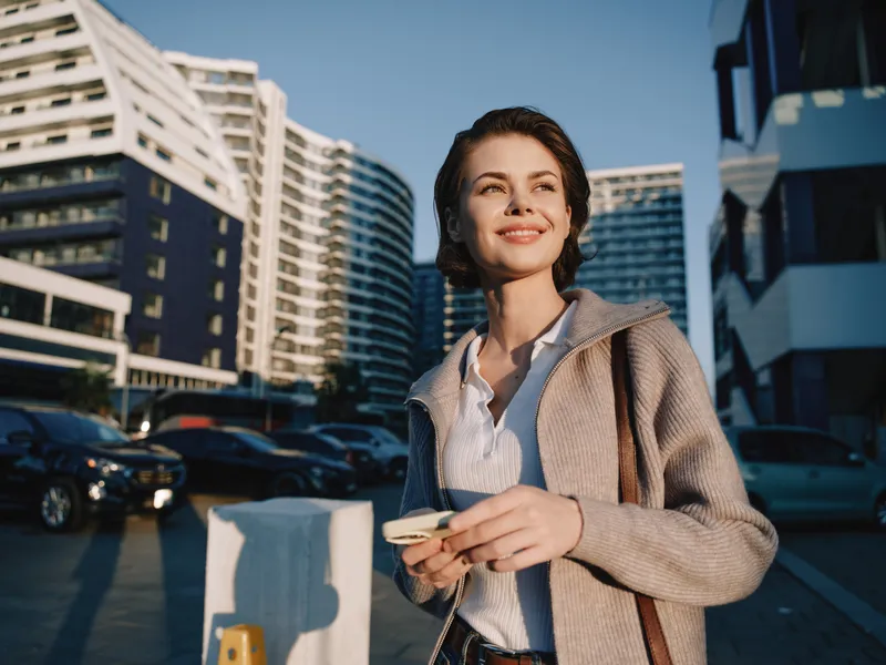 A confident young woman stands outdoors in an urban setting, smiling as she checks her phone, wearing a light cardigan and blouse, with modern buildings in the background.