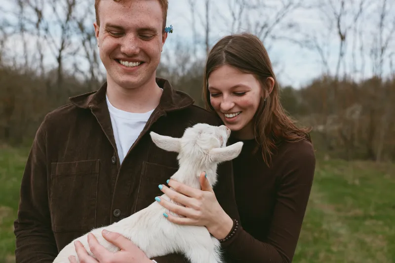 Couple enjoying time with a gentle goat kid outdoors, smiles warm, hands cradle the small animal, park and trees in the background, lighthearted rural moment, shared happiness and bonding with animals