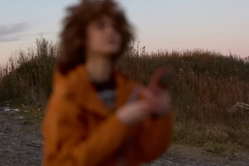 Woman lifestyle outdoors in field at sunset, blurred tall grass and path, woman in warm coat, soft bokeh light, calm nature mood, cinematic portrait