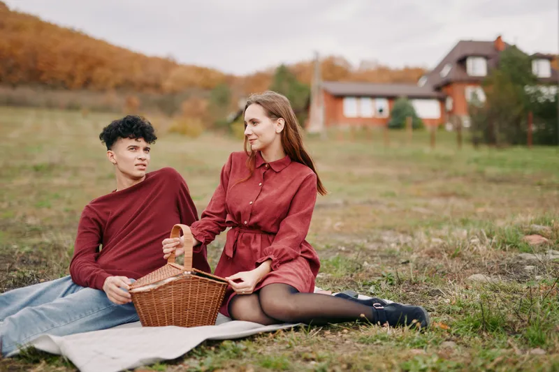 Candid young couple enjoying a countryside picnic on a blanket, sharing a wicker basket and warm eye contact that conveys authentic connection and genuine everyday moments outdoors.