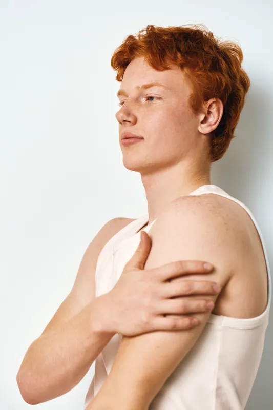 Young man with red hair and pale skin wearing white tank top touching shoulder in studio. Side view with neutral expression and clean background for portrait or health concept.