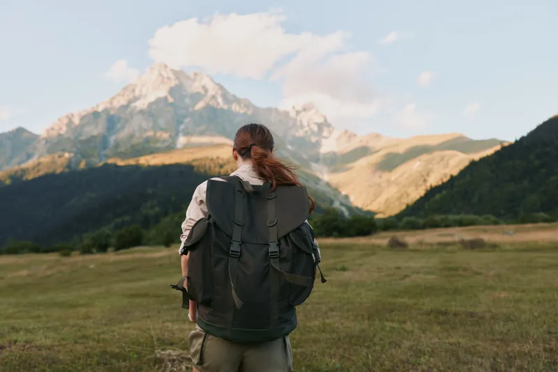 Young woman with a backpack stands in a sunlit mountain meadow, taking in the distant peaks with a calm, focused expression, enjoying the outdoors.