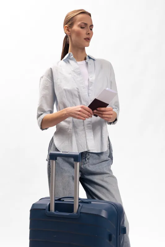 Young woman in casual clothes holds passport and boarding pass, standing with blue suitcase on white background. Concept for travel, vacation, and airport preparation.