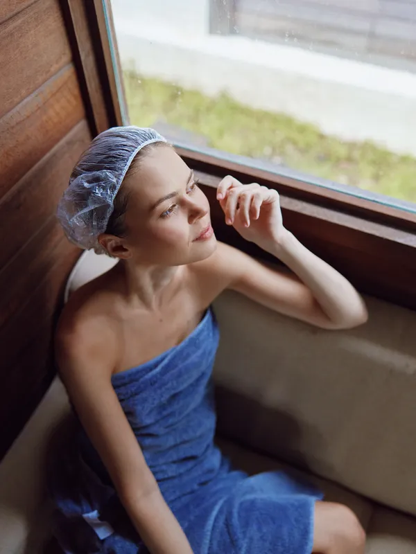 Relaxed woman wrapped in blue towel with hair cover sitting by the window in wooden spa room, enjoying peaceful moment and natural light during wellness treatment.