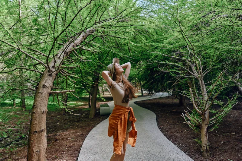 Woman walking on a winding forest path under dense green trees, nature summer scene with flowing dress and relaxed pose, outdoors lifestyle and peaceful woodland pathway.