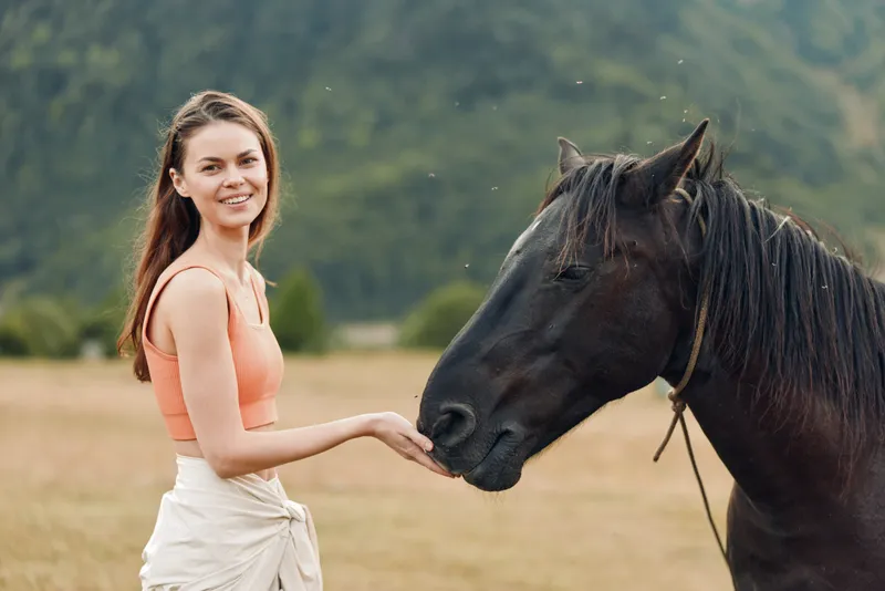 A cheerful woman stands in a grassy field, gently touching a calm horse's nose. She smiles warmly, enjoying a peaceful, rural moment with nature.