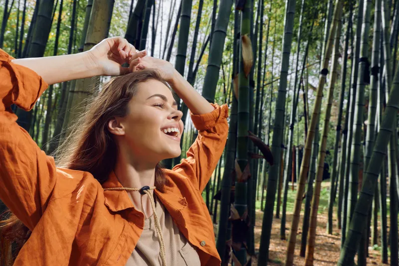 Woman in bamboo forest smiling with eyes closed, portrait of happiness and relaxation outdoors. Young woman in orange jacket enjoying nature and sunlight among tall bamboo stalks.