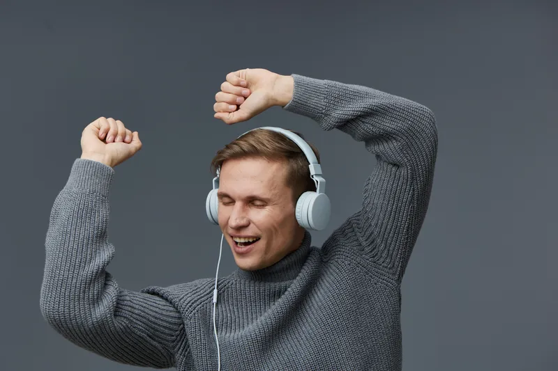 Smiling man with headphones enjoying music or a podcast, raising arms in excitement, casual gray sweater, isolated on plain gray background, lifestyle concept, positive energy, leisure time, entertain
