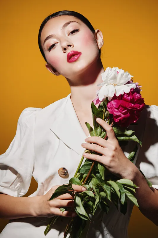 Young woman with glossy red lipstick poses holding bold pink and white flowers, wearing a stylish white blazer against yellow background. Concept for creative beauty fashion.