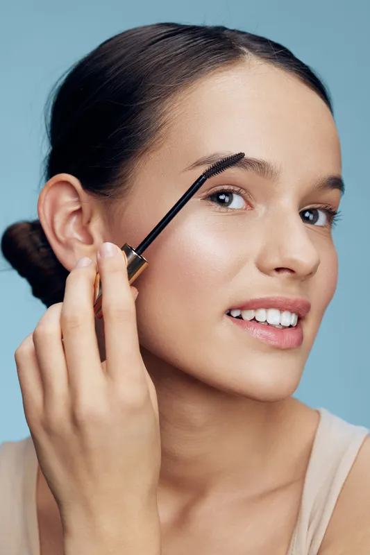 Young woman applying brow gel with a cheerful expression, showcasing beauty routine and natural makeup in soft blue background