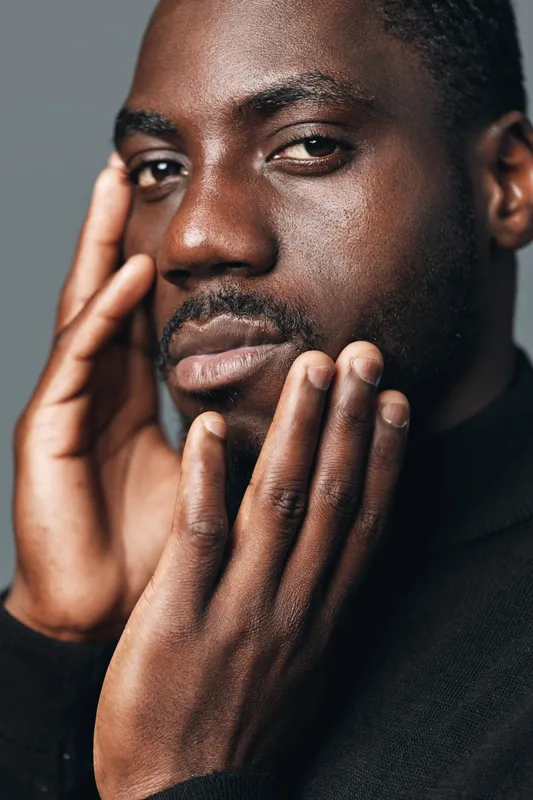 African man with dark skin and short hair touching face in creative conceptual visual style. Closeup portrait with thoughtful expression and natural lighting on black background.