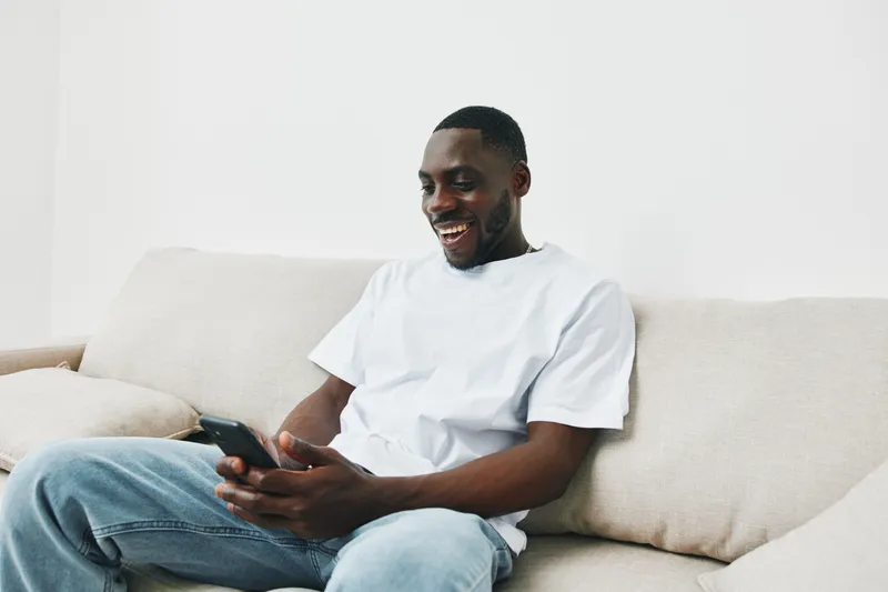 Happy young African American man smiling while using smartphone on a cozy beige sofa, showcasing relaxation and modern lifestyle